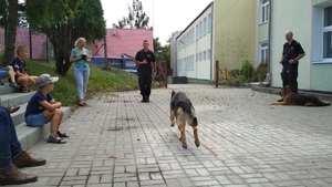na pierwszym planie pies biegnący do swojego policyjnego opiekuna, po lewej stronie uczestnicy spotkania, po prawej drugi z przewodników ze swoim psem