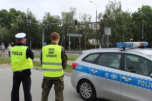 umundurowani policjant i żołnierz WOT stoją przy oznakowanym radiowozu Policji nadzorując bezpieczeństwo w rejonie skrzyżowania i przejścia dla pieszych